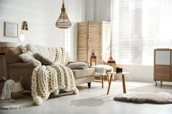 Przytulny salon w stylu hygge wnętrza. Beżowa kanapa z grubym, plecionym kocem, wiklinowa lampa, parawan i świece tworzą ciepłą atmosferę.