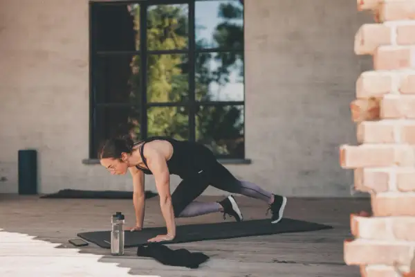 Femme en position de planche, genou vers poitrine, effectuant un exercice polyarticulaire. Bouteille d'eau et téléphone à proximité.