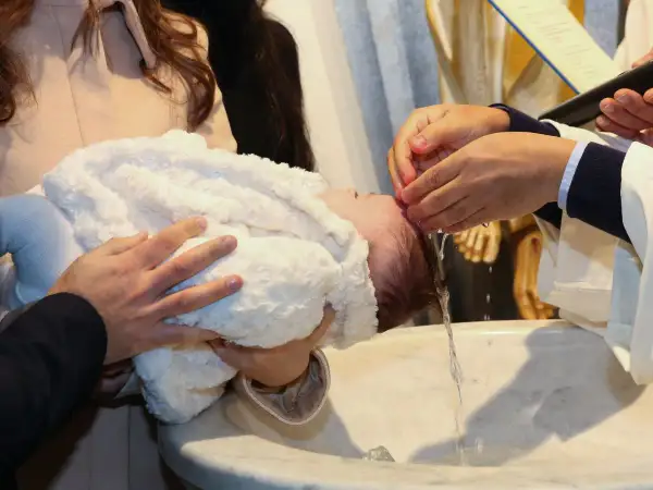 Chrzest święty: ksiądz polewa głowę dziecka wodą. Rodzice trzymają maluszka w białym kocyku.