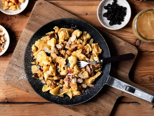 Kaiserschmarrn-Rezepte: Ein goldbrauner Kaiserschmarrn mit Mandelsplittern, Rosinen und Apfelstücken in einer Pfanne, bestäubt mit Puderzucker.