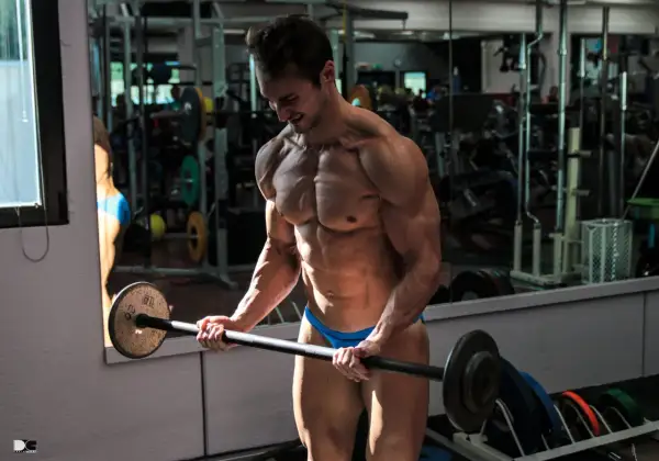 Atleta in palestra, con bicipite e tricipite scolpiti, solleva un bilanciere. Riflessi nello specchio mostrano attrezzature sportive.