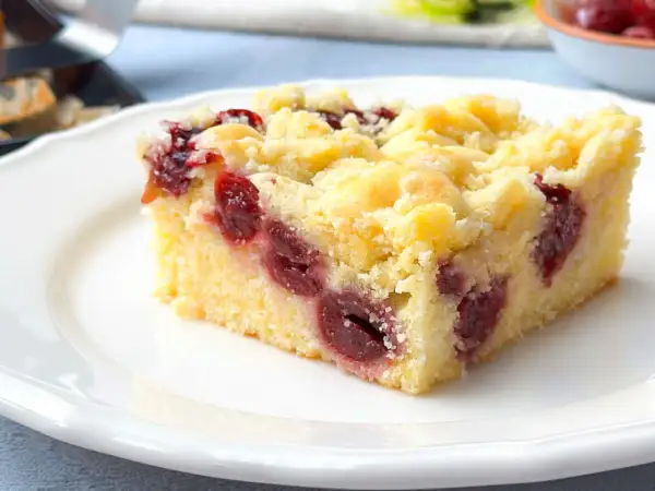 Ein Stück saftiger Kirschstreuselkuchen vom Blech auf einem weißen Teller, mit vielen Kirschen und knusprigen Streuseln.