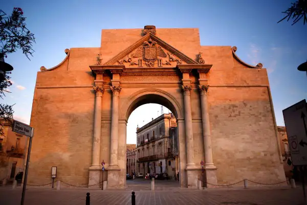 Porta Napoli a Lecce, un maestoso arco in pietra illuminato dal tramonto, accoglie i visitatori con la sua architettura barocca.