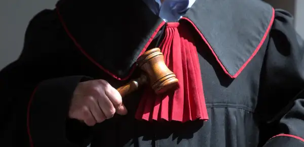 Sędzia trzyma młotek, gotowy do ogłoszenia wyroku. W tle widać togę z czerwonym żabotem, symbolizującą sprawiedliwość i powagę, która towarzyszy wydaniu subsydiarnego aktu oskarżenia.