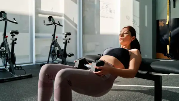 Femme enceinte effectuant un hip thrust au sol avec haltères, concentrée sur son entraînement dans une salle de sport lumineuse.