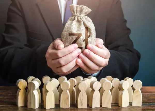 Biznesmen w garniturze trzyma worek z pieniędzmi dla grupy drewnianych ludzików. To symbol, jak siła wyższa może wpływać na losy wielu.