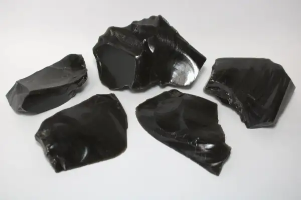 Fünf scharfkantige Stücke glänzenden, schwarzen Obsidiansteins liegen auf weißem Untergrund.