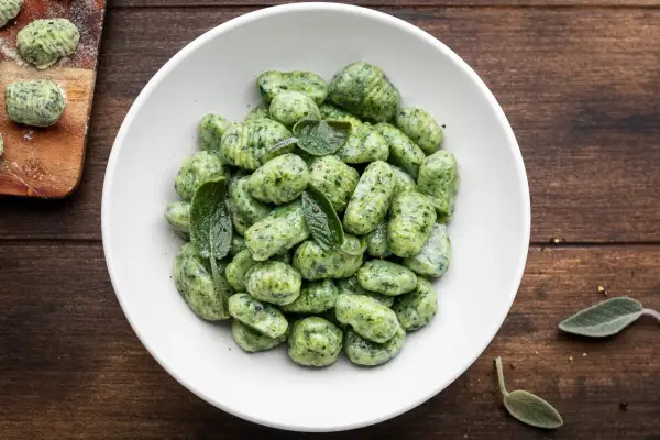 Gnocchi di spinaci freschi, conditi con foglie di salvia, in una ciotola bianca su un tavolo di legno scuro.