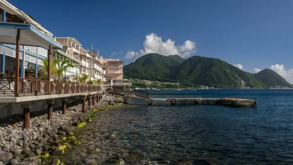 Bâtiments colorés en bord de mer sur l'île Dominique, avec des montagnes verdoyantes en arrière-plan et un quai s'avançant dans l'eau bleue.