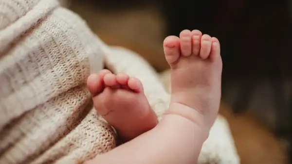 Delikatne, różowe stópki niemowlaka, z widocznymi podwiniętymi palcami u nóg, spoczywają na miękkim, beżowym kocyku.