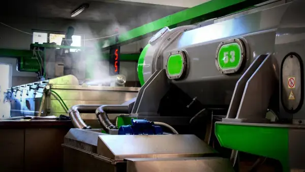 Macchinari industriali in una fabbrica, con tubi e vapore. Si intravede un cartello con "53", forse legato alla produzione di olio di sansa cos'è.