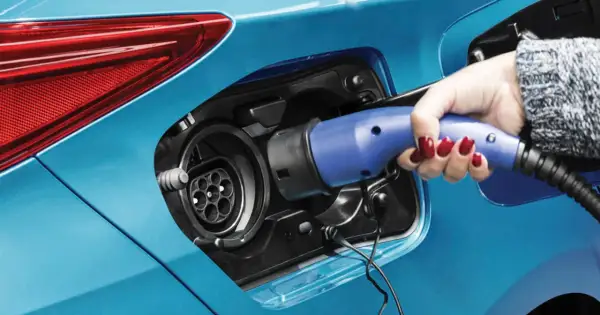 Dłoń z czerwonym lakierem na paznokciach wkłada niebieską wtyczkę do gniazda ładowania samochodu. To przyszłość motoryzacji – hybryda plug-in gotowa do jazdy.