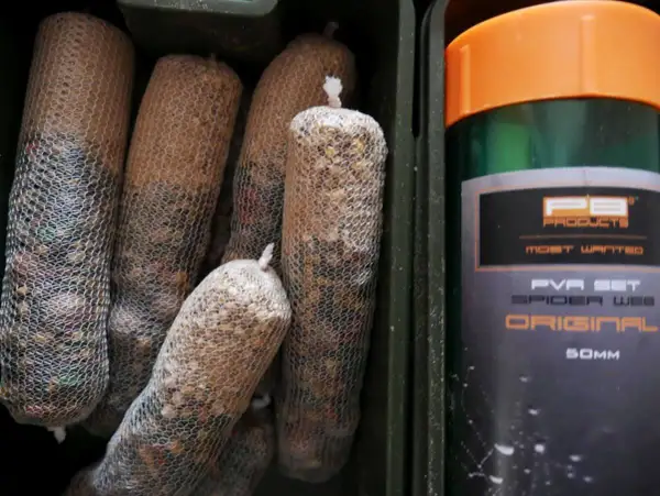 Kilka woreczków z zanętą w siatce PVA, obok zielony pojemnik z pomarańczową nakrętką, na którym widnieje napis "PVA SET SPIDER WEB ORIGINAL".