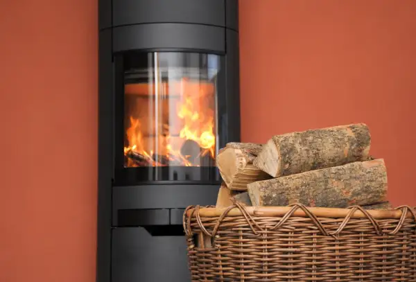Moderner Kaminofen mit brennendem Holz. Ein Korb mit Holzscheiten daneben. Erneuerbare Energien, 15 Prozent Holz als Brennstoff.