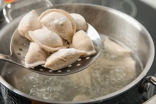 Gotowanie pierogów: na łyżce cedzakowej świeżo uformowane pierogi czekają na wrzucenie do wrzątku. W garnku bulgocze woda, gotowe do przyjęcia pysznego farszu.