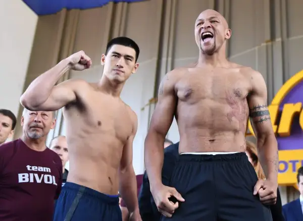 Two boxers face off. One, wearing a "team BIVOL" shirt, flexes his bicep. The other, with tattoos, shouts. Bivol fights in the light heavyweight class.