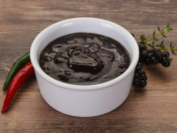 Gęsty, ciemny sos pieprzowy w białej miseczce, obok czerwona i zielona papryczka chili oraz gałązka jeżyn.