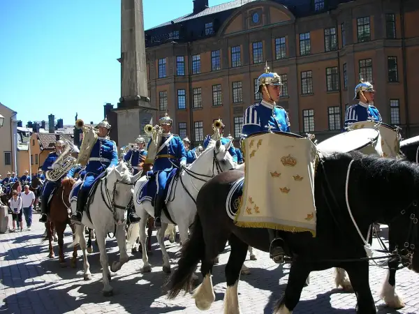 Livgardets dragonmusikkår marscherar i parad. Musiker i blå uniformer spelar på instrument och rider på vita hästar.