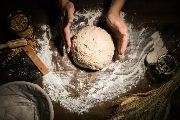 Hände formen einen Teigball auf einer mit Mehl bestäubten Holzoberfläche. Körner, ein Schaber und ein Gärkorb liegen daneben.