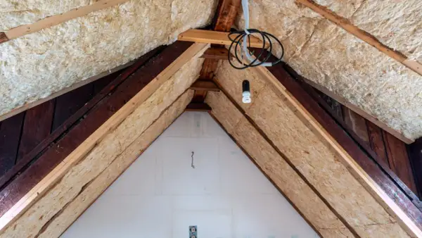 Dämmmaterial wird in einem Dachboden verlegt, um das Haus zu isolieren. Holzbalken und Kabel sind sichtbar.