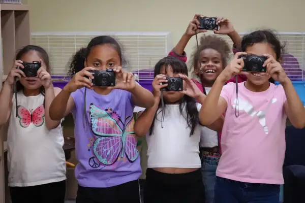 Five girls hold up old cameras, learning what to do with them by taking pictures.