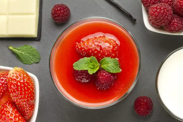 Un bicchiere di coulis di fragole con fragola, lamponi e menta. Accanto, fragole fresche, cioccolato bianco, latte e un baccello di vaniglia.