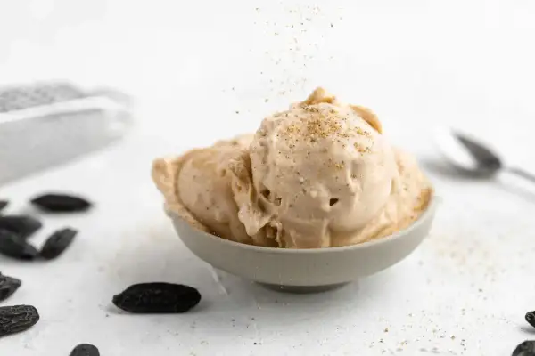 Schüssel mit cremigem Tonka Eis, bestreut mit gemahlenen Tonkabohnen. Dunkle Tonkabohnen liegen verstreut auf dem Tisch.