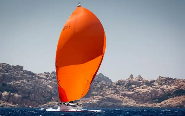 Vela arancione brillante, un gennaker avvolgibile, gonfia al vento su un'imbarcazione a vela che naviga vicino a una costa rocciosa.