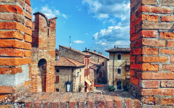 Un antico borgo vicino Parma, con case in pietra e tetti rossi, incorniciato da mura di mattoni sotto un cielo azzurro e nuvoloso.