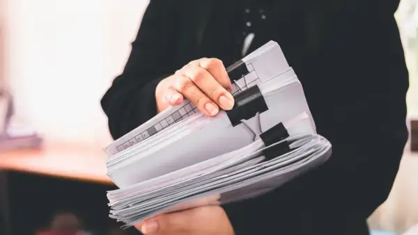 Osoba w czerni trzyma stos dokumentów, wśród których znajduje się ewidencja przebiegu pojazdu dla celów VAT.