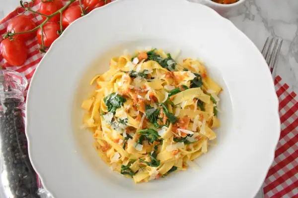 Pyszny makaron ze szpinakiem i pomidorami, posypany parmezanem. Obok świeże pomidorki i młynek do pieprzu.