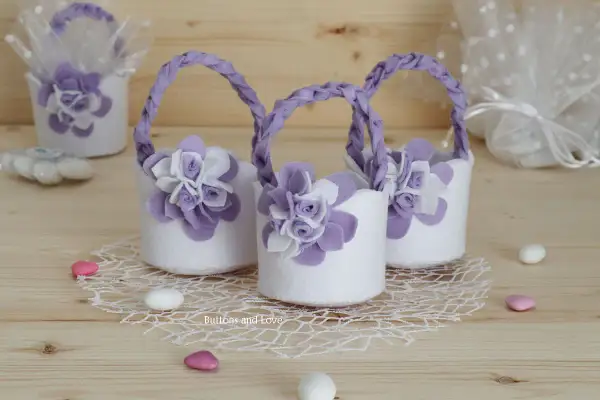 Piccoli cestini porta bomboniere fai da te in feltro bianco con fiori lilla e manici intrecciati, perfetti per un evento speciale.