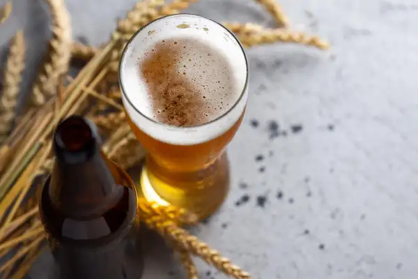 Bicchiere di birra di frumento dorata con schiuma cremosa, accanto a una bottiglia scura e spighe di grano su sfondo grigio.