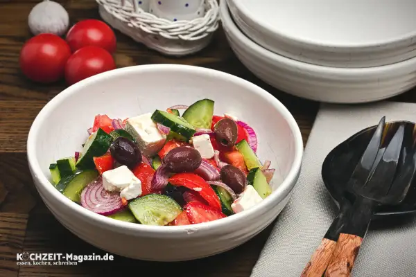 Frischer griechischer Salat mit Tomaten, Gurken, roten Zwiebeln, Oliven und Feta in einer weißen Schüssel.