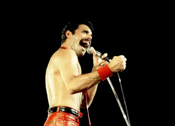 Freddie Mercury w czerwonych spodniach i opasce na nadgarstku śpiewa do mikrofonu. Nawet jako dorosły, jego energia przypominała dzikie dziecięce zabawy.