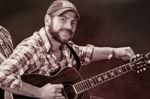Kyle Shearer, a bearded man in a plaid shirt and cap, strums his acoustic guitar, ready to play a tune.