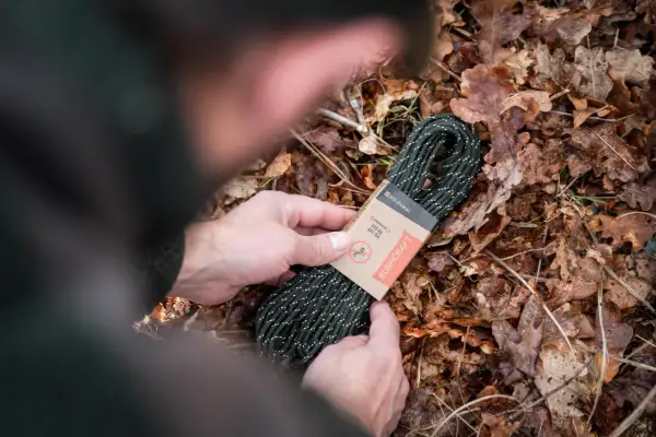 Manos sostienen un rollo de cordón paracord verde con puntos blancos, sobre un lecho de hojas secas.
