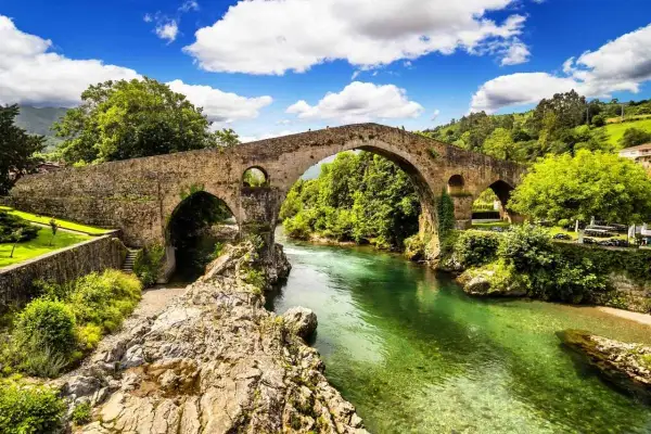 Majestuoso puente romano asturias sobre un río de aguas cristalinas, rodeado de exuberante vegetación y un cielo azul con nubes blancas.