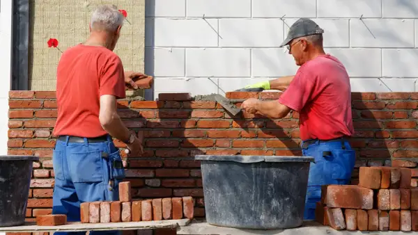 Dwóch budowlańców stosuje zasady murowania z cegły pełnej, układając cegły na zaprawie.