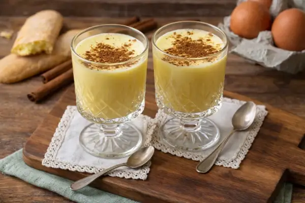 Zwei Gläser cremiges Zabaione ohne Alkohol, bestreut mit Zimt. Dazu Savoiardi-Kekse und Zimtstangen.