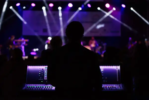 Silueta de técnico de sonido operando una mesa de mezclas en un plano de escenario musical, con luces púrpuras y blancas iluminando la banda al fondo.