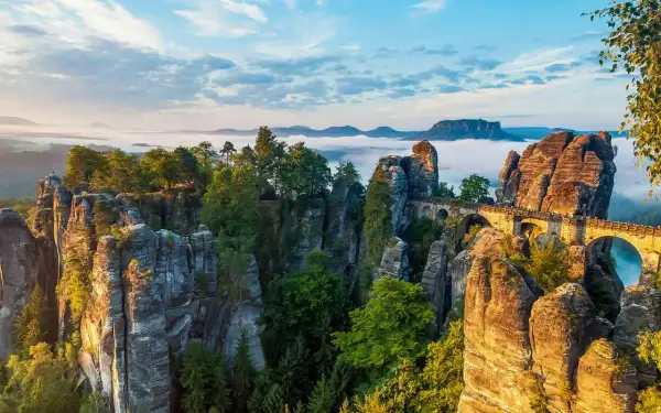 Felsformationen und eine Basteibrücke in der Böhmischen Schweiz, umgeben von Nebel und Bäumen, unter einem blauen Himmel.
