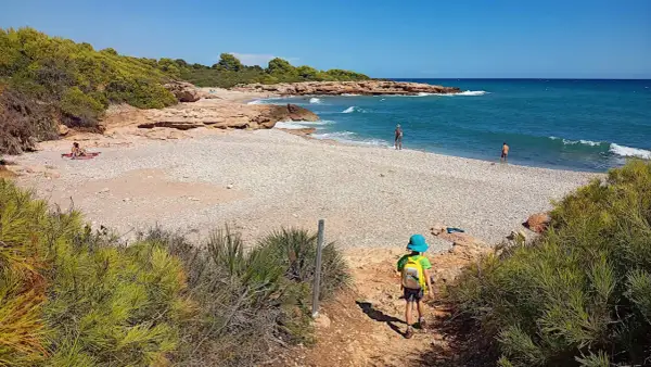 Niño con mochila azul desciende por sendero hacia la playa de rueta, con rocas y mar turquesa.