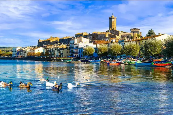 Anatre nuotano in un lago sereno, con un borgo pittoresco e barche colorate che si specchiano sull'acqua. Un'immagine da cartolina dei laghi italiani.
