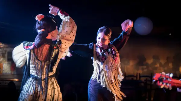 Dos bailarinas de flamenco, con trajes de lunares y flecos, interpretan apasionadas canciones de sevillanas bajo luces de escenario.