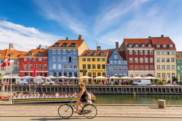 En kvinna cyklar längs Nyhavn i Köpenhamn, ett soligt exempel på danmark klimat. Färgglada hus och kanalbåtar skapar en livlig atmosfär.