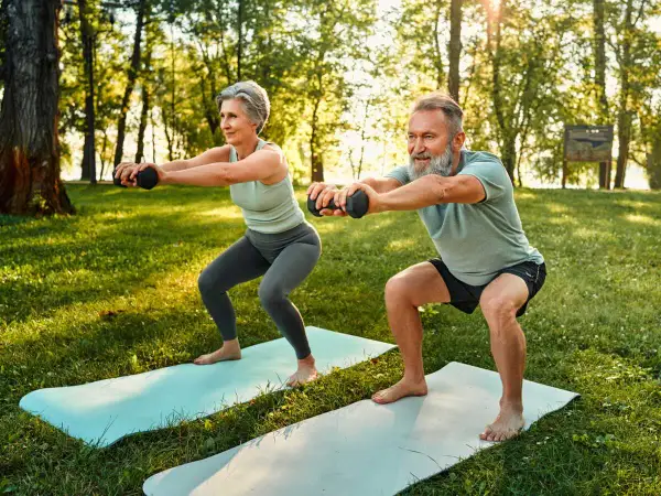Une femme sportive de 50 ans et un homme font des squats avec des haltères dans un parc ensoleillé.