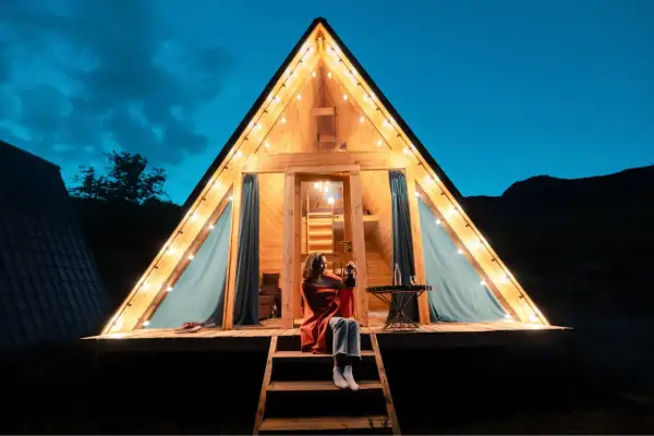 Soirée de camping nature corse. Une femme assise sur les marches d'un chalet en bois illuminé par des guirlandes lumineuses, profitant de la nuit.
