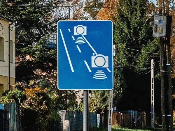 Niebieski znak drogowy z białymi symbolami aparatów fotograficznych i falami, informujący o odcinkowym pomiarze prędkości.