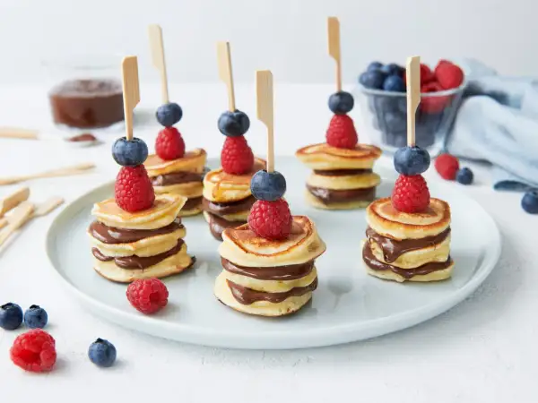Mini pancakes rezept: gestapelte Mini-Pfannkuchen mit Schokocreme, garniert mit Himbeeren und Blaubeeren auf Spießen.